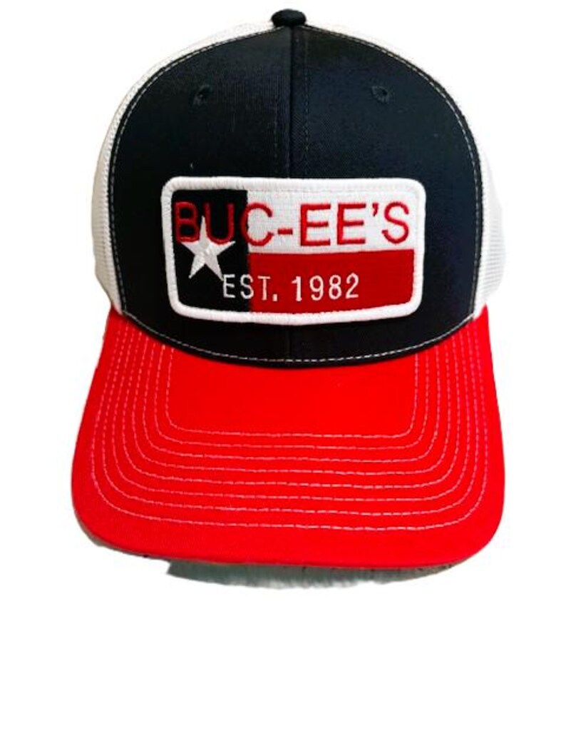 May include: A black, red, and white baseball cap with a mesh back. The front features a rectangular patch with the text "BUC-EE'S" in red letters, a white star, and the text "EST. 1982". The brim is red.