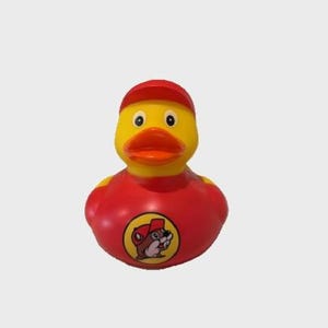May include: A yellow rubber duck wearing a red cap and shirt. The duck has an orange beak and a logo of a beaver wearing a cap on its shirt. The duck is a novelty item.