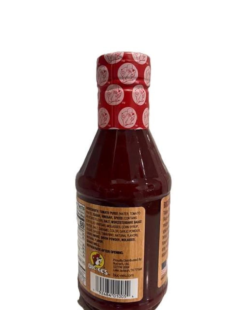 May include: A bottle of Buc-ee's brand sauce with a red cap featuring a repeating logo pattern. The bottle is filled with a dark red liquid and has a label with ingredient information. The label also includes the Buc-ee's logo.