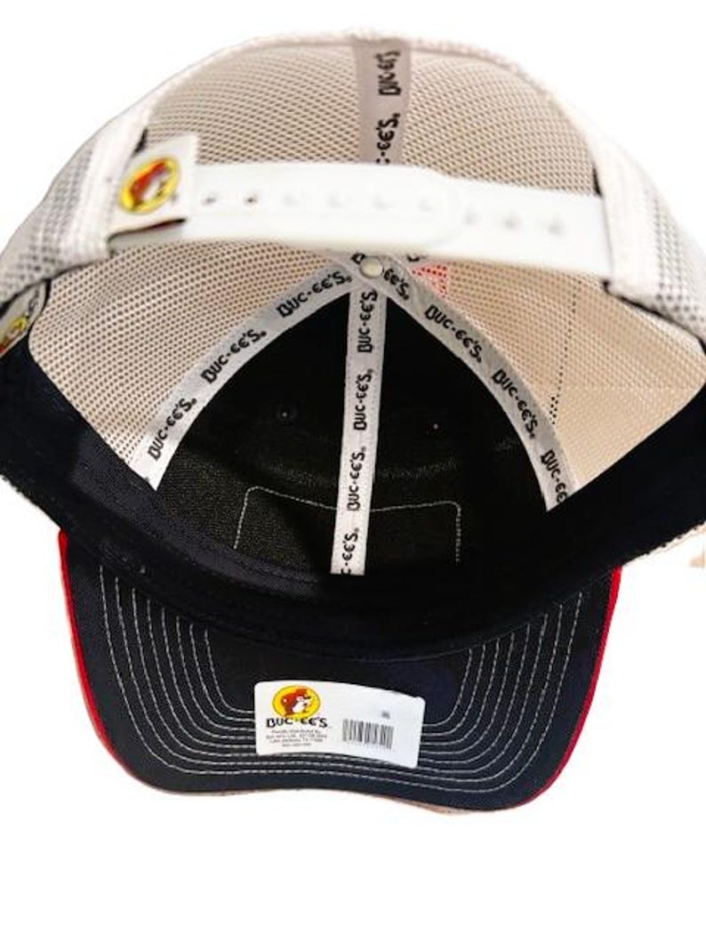 May include: A navy blue and red baseball cap with a white mesh back. The cap features the "Buc-ee's" logo and text on the inside and a white adjustable strap. The brim is navy blue with red trim.