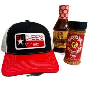 May include: A black and red baseball cap with the text "BUC-EE'S EST. 1982" on the front, along with a bottle of BBQ sauce and a jar of BBQ rub. The cap has a white mesh back and a red brim.