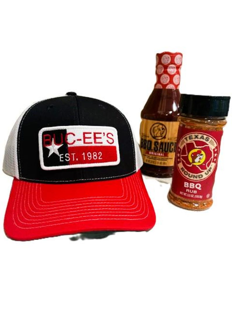 May include: A black and red baseball cap with the text "BUC-EE'S EST. 1982" on the front, along with a bottle of BBQ sauce and a jar of BBQ rub. The cap has a white mesh back and a red brim.
