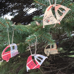 May include: Four paper ornaments shaped like campers and sailboats hang from a pine tree branch. The ornaments are decorated with red, white, and blue paper and have a vintage look.