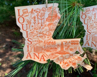 May include: A wooden ornament shaped like the state of Louisiana with the words "LOUISIANA" printed on it. The ornament is decorated with orange illustrations of various Louisiana landmarks and symbols, including a steamboat, a crawfish, and a fleur-de-lis.