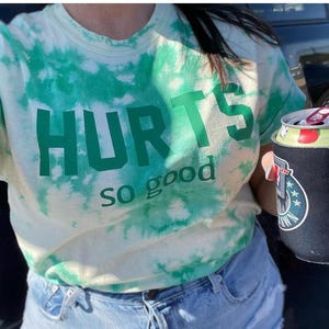 Puede incluir: Una camiseta blanca de tie-dye con texto verde que dice "HURTS so good".