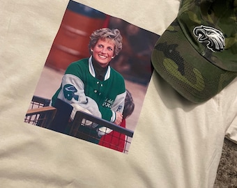 May include: A white t-shirt with a printed image of a woman wearing a green and white jacket. The woman is smiling and looking at the camera. The image is centered on the shirt.