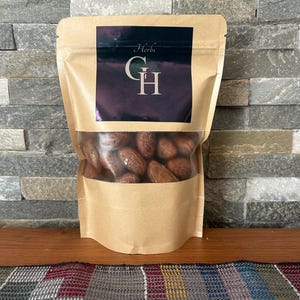 以下が含まれることがあります： 透明な窓が付いた茶色の紙袋に、乾燥した茶色のナッツが入っています。袋には黒いラベルが付いており、"Herbs"という文字と、スタイリッシュなフォントで"GH"という文字が書かれています。