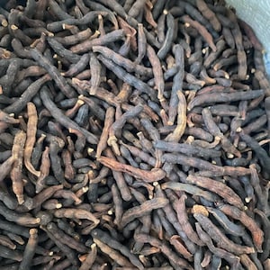 May include: A close-up of a heap of dried, dark brown, elongated spices. The spices have a wrinkled texture and a slightly curved shape. The overall colour palette is dominated by shades of brown and black.