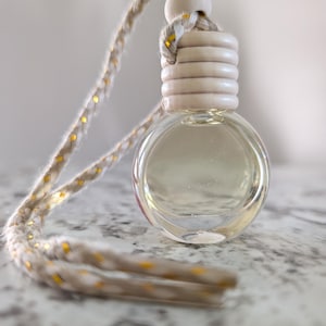 Puede incluir: Un frasco pequeño de vidrio transparente y forma redonda, lleno de un líquido claro y coronado con un tapón de madera estriado. Un cordón beige trenzado con motas doradas metálicas está sujeto al tapón para colgar el frasco.