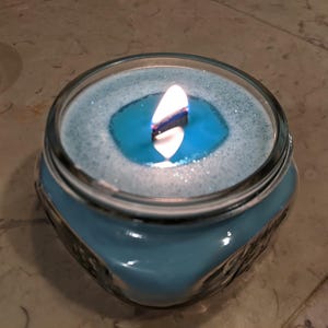 May include: A blue glass candle jar with a white wax top and a single flame burning in the center. The wax has a blue tint and a white layer on top.