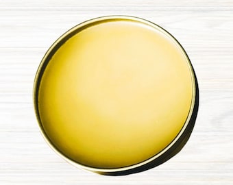 May include: A round, gold-coloured tin container filled with a smooth, yellow substance.