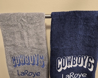 May include: Two embroidered hand towels. One is gray with blue embroidery reading "COWBOYS" and "LaRoye." The other is navy blue with white embroidery reading "COWBOYS" and "LaRoye." Both towels are hanging on a metal bar.