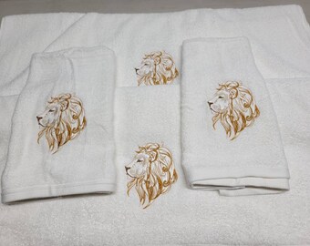 May include: Set of four white towels with embroidered lion head designs in gold. The towels are folded and arranged on a white surface.
