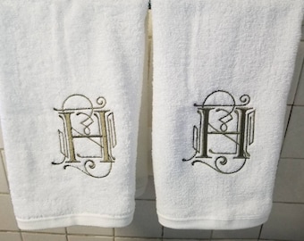 May include: Two white bath towels with an embroidered gold and dark brown letter "H" design. The towels are hanging on a metal bar, showcasing the detailed monogram. The towels are made of a soft, absorbent material.