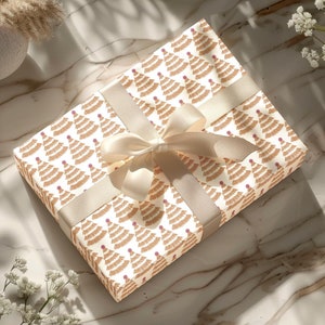 May include: A gift box wrapped in white paper with a brown and pink Christmas tree pattern. The box is tied with a cream-colored ribbon.