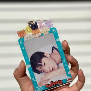 May include: A turquoise blue plastic photo card holder with a clear plastic photo insert. The holder has a white border with a cartoon ship's wheel, musical notes, and stars. There are eight small cartoon animal charms on the top of the holder. The photo insert shows a young man with dark hair lying down. The text "Ateez" is printed on the bottom of the holder.