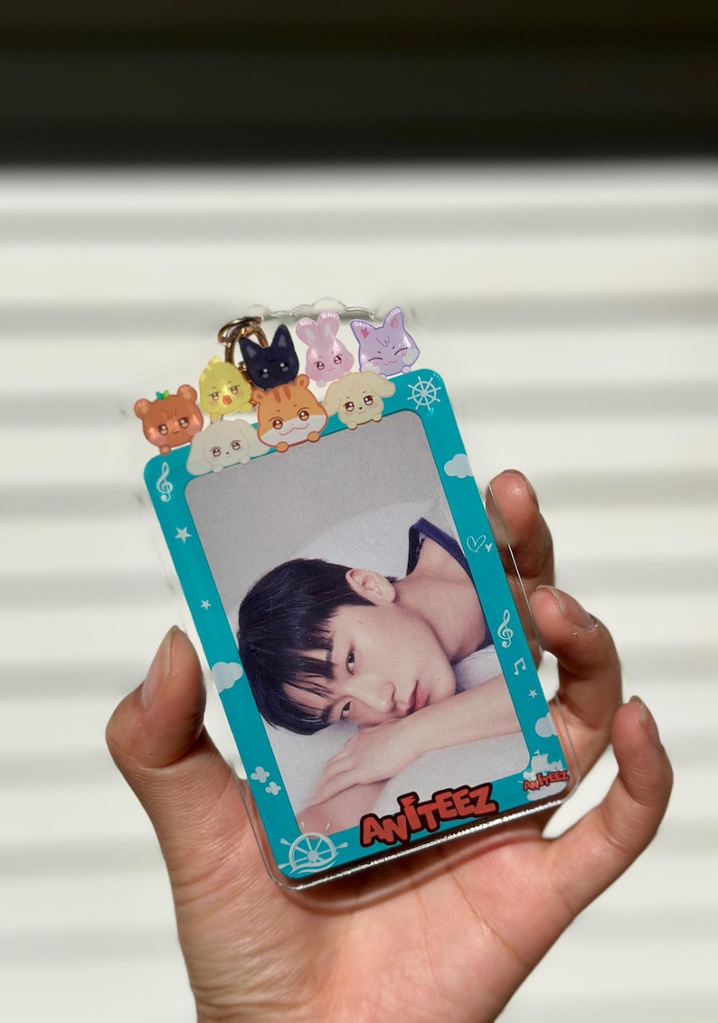 May include: A turquoise blue plastic photo card holder with a clear plastic photo insert. The holder has a white border with a cartoon ship's wheel, musical notes, and stars. There are eight small cartoon animal charms on the top of the holder. The photo insert shows a young man with dark hair lying down. The text "Ateez" is printed on the bottom of the holder.