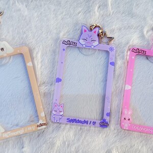 May include: Three clear plastic photo card holders with different coloured backgrounds and cartoon animal charms. The holders have a gold star charm attached to the top. The text on the holders reads "Ateez" and the names of the members.