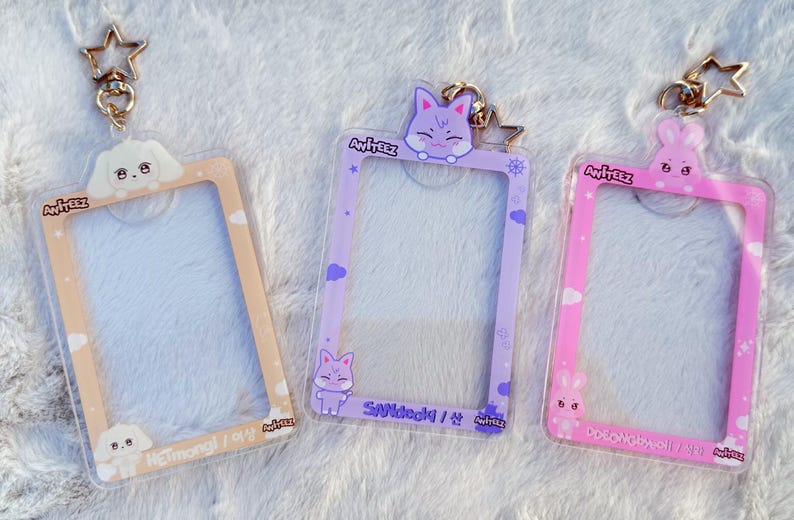 May include: Three clear plastic photo card holders with different coloured backgrounds and cartoon animal charms. The holders have a gold star charm attached to the top. The text on the holders reads "Ateez" and the names of the members.