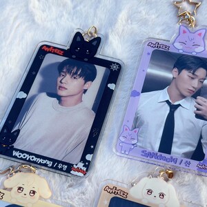 May include: Two clear plastic photo card holders with a black and purple frame. Each holder features a different member of the K-pop group Ateez. The top of each holder has a cartoon animal charm. The black frame holder has a black cat charm and the purple frame holder has a purple cat charm. The text "Ateez" is printed on each holder.