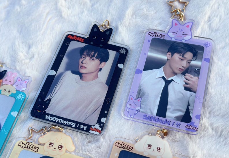 May include: Two clear plastic photo card holders with a black and purple frame. Each holder features a different member of the K-pop group Ateez. The top of each holder has a cartoon animal charm. The black frame holder has a black cat charm and the purple frame holder has a purple cat charm. The text "Ateez" is printed on each holder.