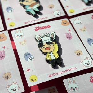 May include: A pink and white enamel pin featuring a cartoon character with a yellow shirt and a white bunny on their head. The pin has the text "Skzoo" and "@afteryouartshop" on it.