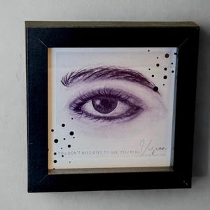 May include: Framed artwork featuring a detailed pencil drawing of an eye and eyebrow. The artwork is in a black frame and includes the text "YOU DON'T NEED EYES TO SEE, YOU NEED Vision". Black dots are scattered around the image.