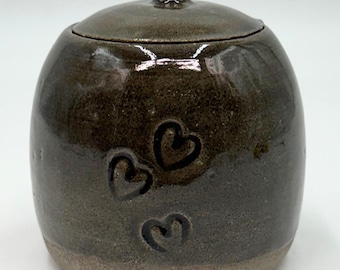 May include: A handmade ceramic jar with a lid. The jar is a dark brown color with three heart-shaped designs. The jar has a rounded shape and a small knob on the lid. The base of the jar is a lighter brown color.