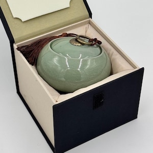 May include: A celadon green ceramic urn with a lid and a bronze-colored ring, presented in a black and beige box. The urn has a rounded shape and a tassel. The box is open, revealing the urn nestled inside.