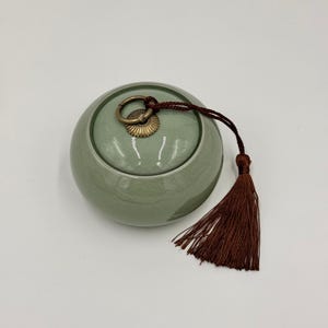 May include: A round, sage green ceramic jar with a lid, featuring a brass handle and a decorative brown tassel. The jar has a smooth, glossy finish and a crackle glaze effect, suggesting a vintage or handcrafted aesthetic.
