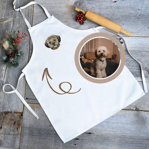 May include: White apron with a brown embroidered dog face and a circular photo of a dog. A brown arrow points to the photo. A wooden rolling pin and holiday decorations are in the background. The apron has white ties.