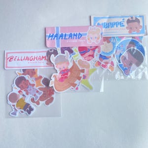 May include: Three clear plastic sticker packs featuring cartoon illustrations of football players. The packs are labelled Bellingham Sticker Pack, Haaland Sticker Pack, and Mbappé Sticker Pack, each containing multiple die-cut stickers of the athletes.