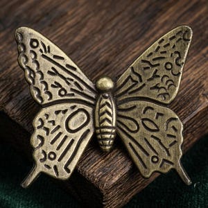 May include: A butterfly-shaped metal pin with an antique brass finish. The wings feature intricate, embossed geometric patterns, including lines, circles, and dashes. The central body is segmented with a chevron design, and the edges are defined.