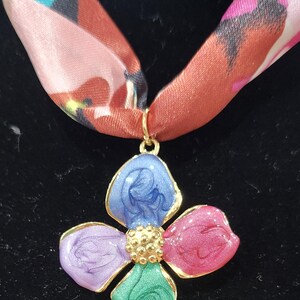 May include: A floral pendant necklace with a colorful design. The pendant is a four-petal flower with blue, pink, green, and purple petals, outlined in gold. The necklace has a patterned silk scarf.