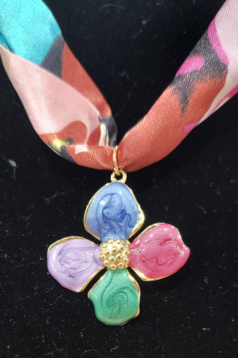 May include: A floral pendant necklace with a colorful design. The pendant is a four-petal flower with blue, pink, green, and purple petals, outlined in gold. The necklace has a patterned silk scarf.