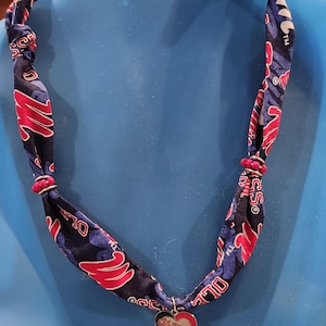 May include: A fabric necklace with a blue and red pattern, featuring a heart-shaped pendant. The pendant has a red heart with the words "Ole Miss" in white. The necklace is displayed on a blue mannequin.