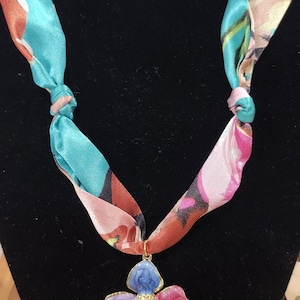 May include: A colorful floral pendant necklace. The pendant is a flower with blue, purple, pink, and green petals. The necklace's ribbon is a mix of teal, pink, and orange. The necklace is displayed on a black velvet surface.