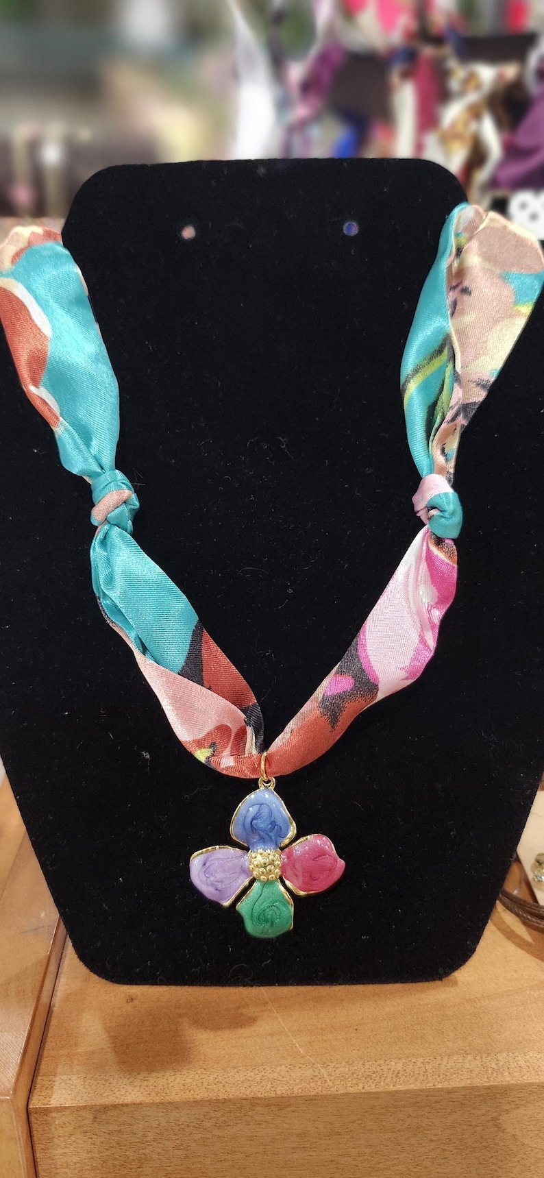 May include: A colorful floral pendant necklace. The pendant is a flower with blue, purple, pink, and green petals. The necklace's ribbon is a mix of teal, pink, and orange. The necklace is displayed on a black velvet surface.
