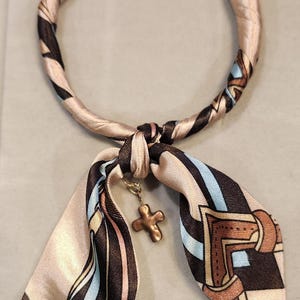 May include: A silk scarf with a repeating pattern of black, blue, and tan stripes and geometric shapes. The scarf is tied in a knot with a gold-colored cross charm hanging from it. The scarf is a fashion accessory.
