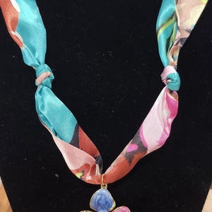 May include: A floral pendant necklace with a colorful design. The pendant is a four-petal flower with blue, purple, pink, and green enamel petals, and a gold-colored center. The necklace's scarf-like straps are teal and pink with a floral pattern.