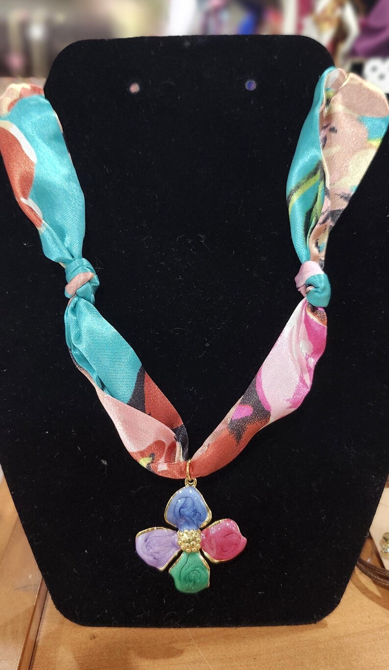 May include: A floral pendant necklace with a colorful design. The pendant is a four-petal flower with blue, purple, pink, and green enamel petals, and a gold-colored center. The necklace's scarf-like straps are teal and pink with a floral pattern.