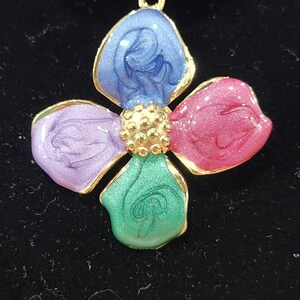 May include: A colorful flower pendant with four petals in blue, pink, green, and purple, each outlined in gold. The center features a cluster of small, gold-colored beads. The pendant is likely made of enamel or a similar material.