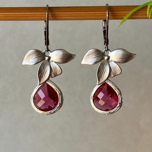以下が含まれることがあります： シルバーカラーのドロップピアス。各イヤリングは上部に花のモチーフがあり、ファセット加工された、涙の形をした、深い赤色の宝石が付いています。イヤリングはレバーバッククロージャーから吊り下げられています。