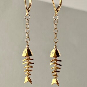 Op de afbeelding: Gouden visgraatoorbellen met een klein diamant accent op elke vis. De oorbellen zijn aan een ketting en hebben een haak sluiting.