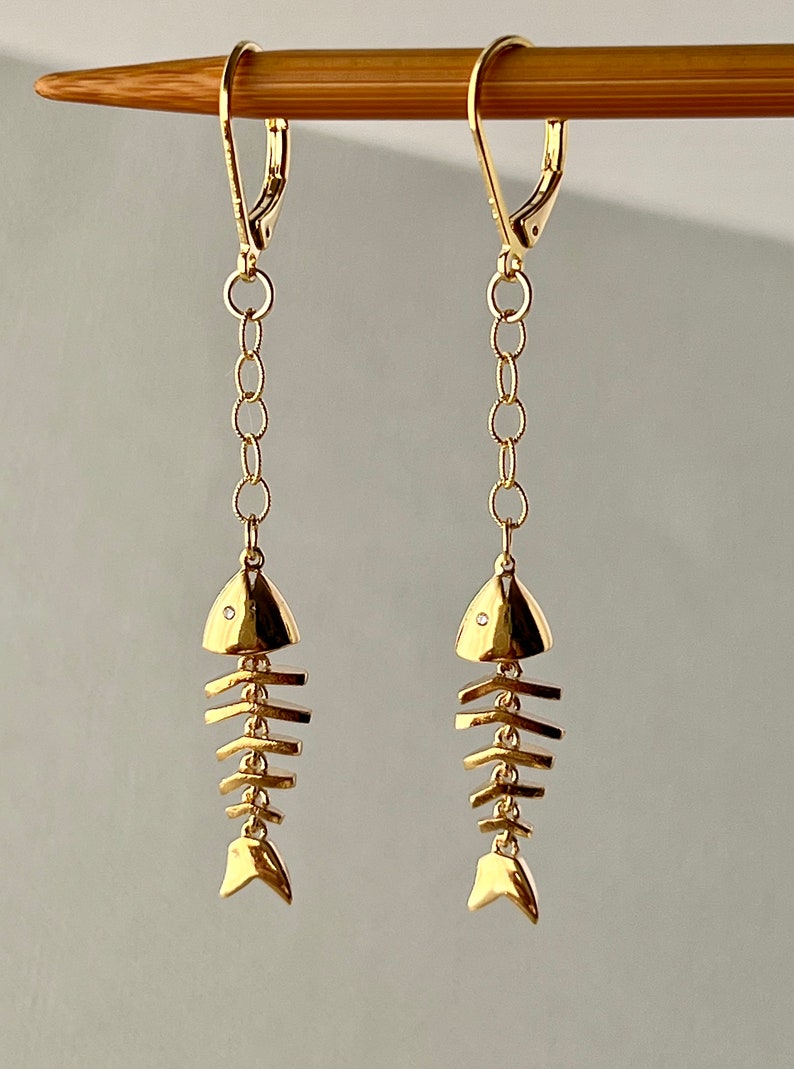 Op de afbeelding: Gouden visgraatoorbellen met een klein diamant accent op elke vis. De oorbellen zijn aan een ketting en hebben een haak sluiting.