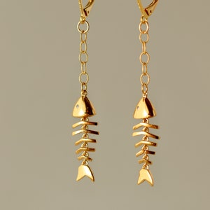 Op de afbeelding: Gouden visgraat oorhangers met een kleine, heldere steen op elke vis. De oorhangers hangen aan een houten stok.