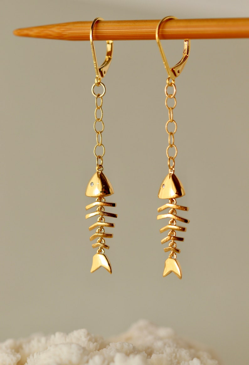 Op de afbeelding: Gouden visgraat oorhangers met een kleine, heldere steen op elke vis. De oorhangers hangen aan een houten stok.