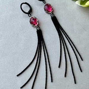 May include: A pair of dangle earrings featuring oval, faceted, ruby-colored gemstones set in silver-toned metal. The earrings have black, looped ear wires and long, black tassel accents, creating a dramatic, elegant look.