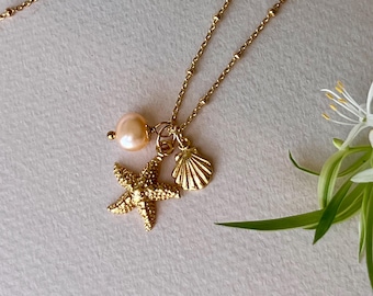 May include: A gold-colored necklace featuring a starfish, seashell, and a peach-colored pearl pendant. The delicate chain is adorned with small, round beads. The necklace is a beach-themed jewelry item.