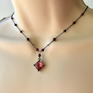 May include: A delicate black chain necklace with small, dark red beads and a square-shaped pendant with a dark red gemstone.